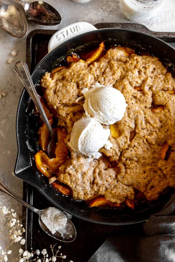 Spoon digging into peach cobbler topped with vanilla ice cream in a cast iron skillet.