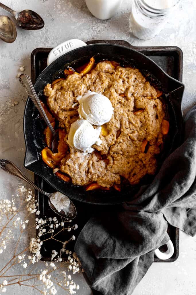 Spoon digging into peach cobbler topped with vanilla ice cream in a cast iron skillet.