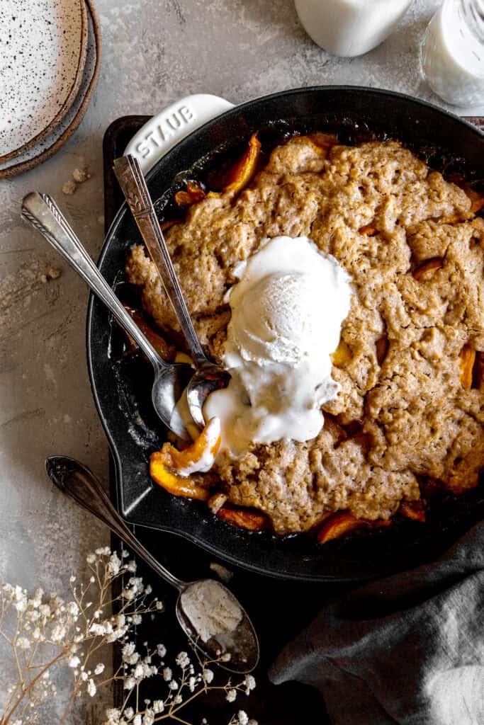 Two spoons digging into peach cobbler and vanilla ice cream in a skillet.