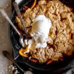 Two spoons digging into peach cobbler and vanilla ice cream in a skillet.