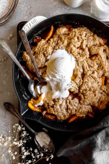 Two spoons digging into peach cobbler and vanilla ice cream in a skillet.