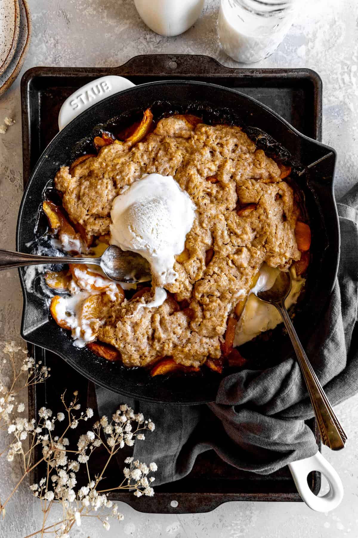 Two spoons digging into peach cobbler and vanilla ice cream in a skillet.