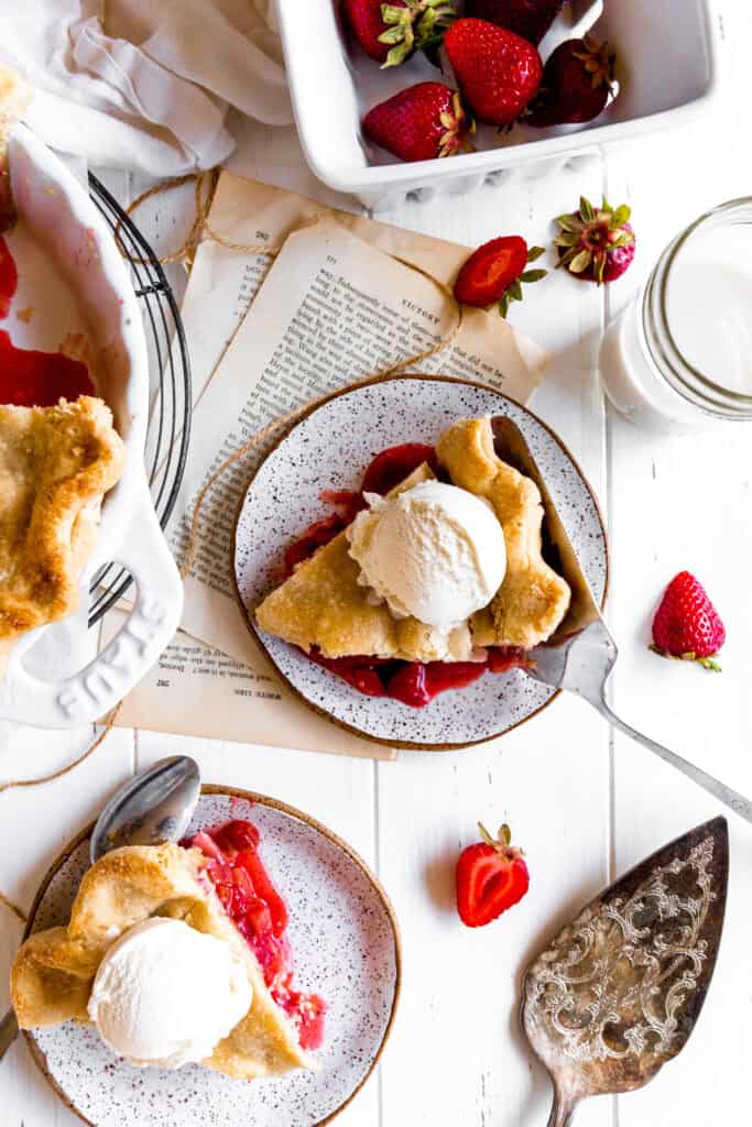 Slices of strawberry rhubarb pie on plates, glasses on milk and scattered berries.