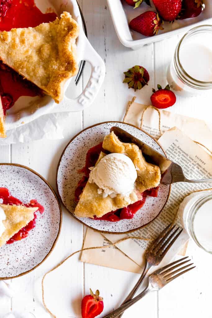 Slices of strawberry rhubarb pie on plates, glasses of milk and a carton of strawberries.
