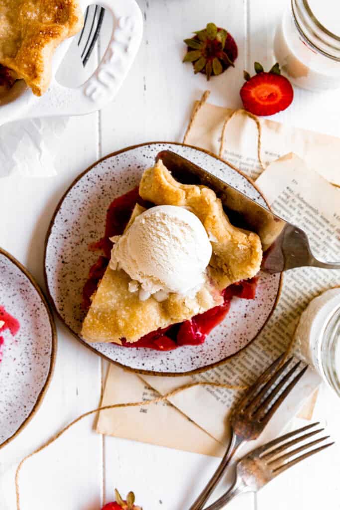 Slice of strawberry rhubarb pie on a plate, a glass of milk and two forks.