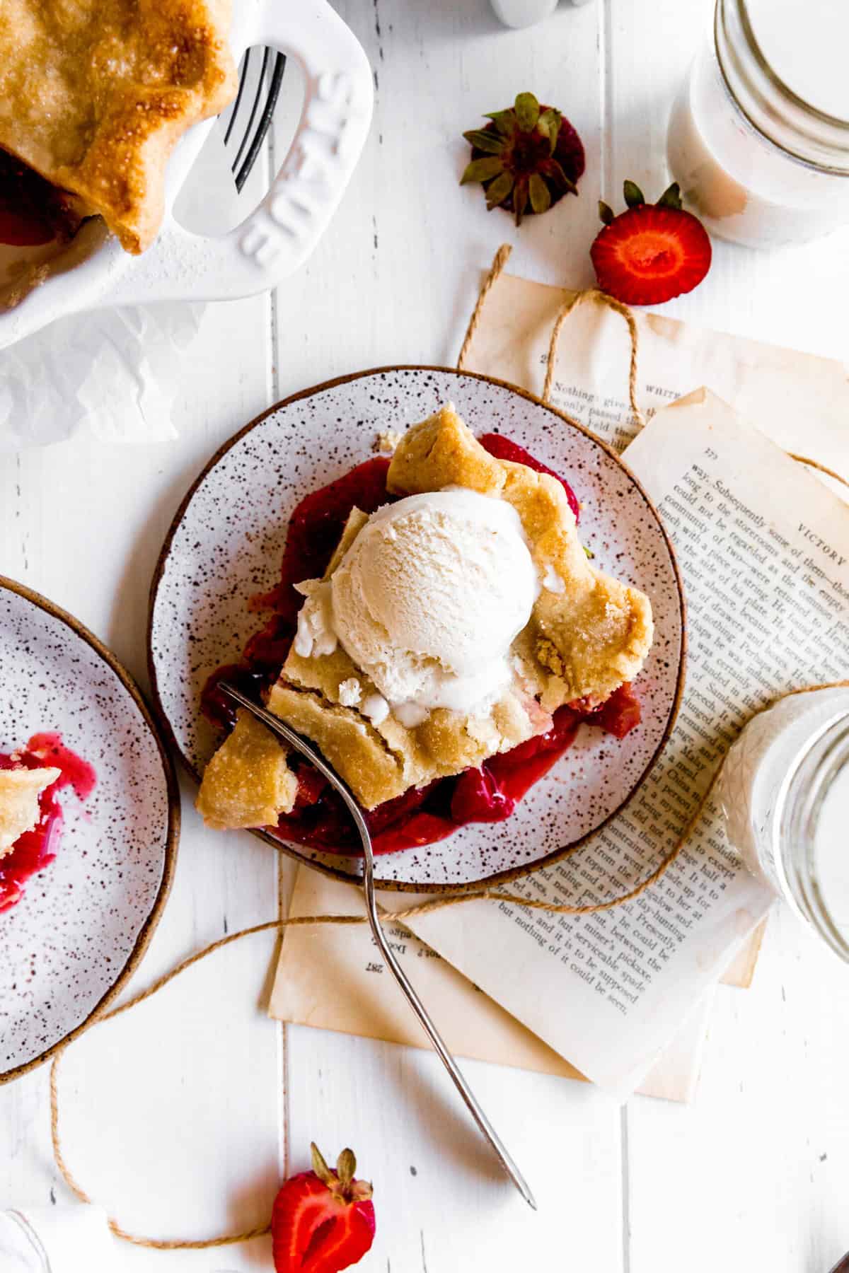 Fork cutting into a slice of strawberry rhubarb pie on a white plate and glasses of milk.