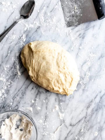 Pizza dough on a marble surface and a bowl of flour.