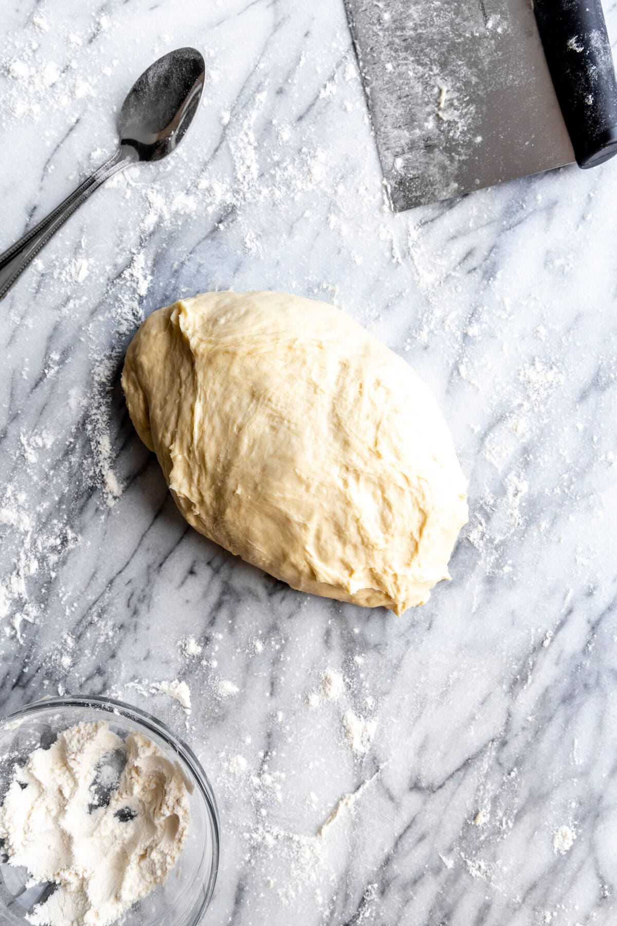 Pizza dough on a marble surface and a bowl of flour.
