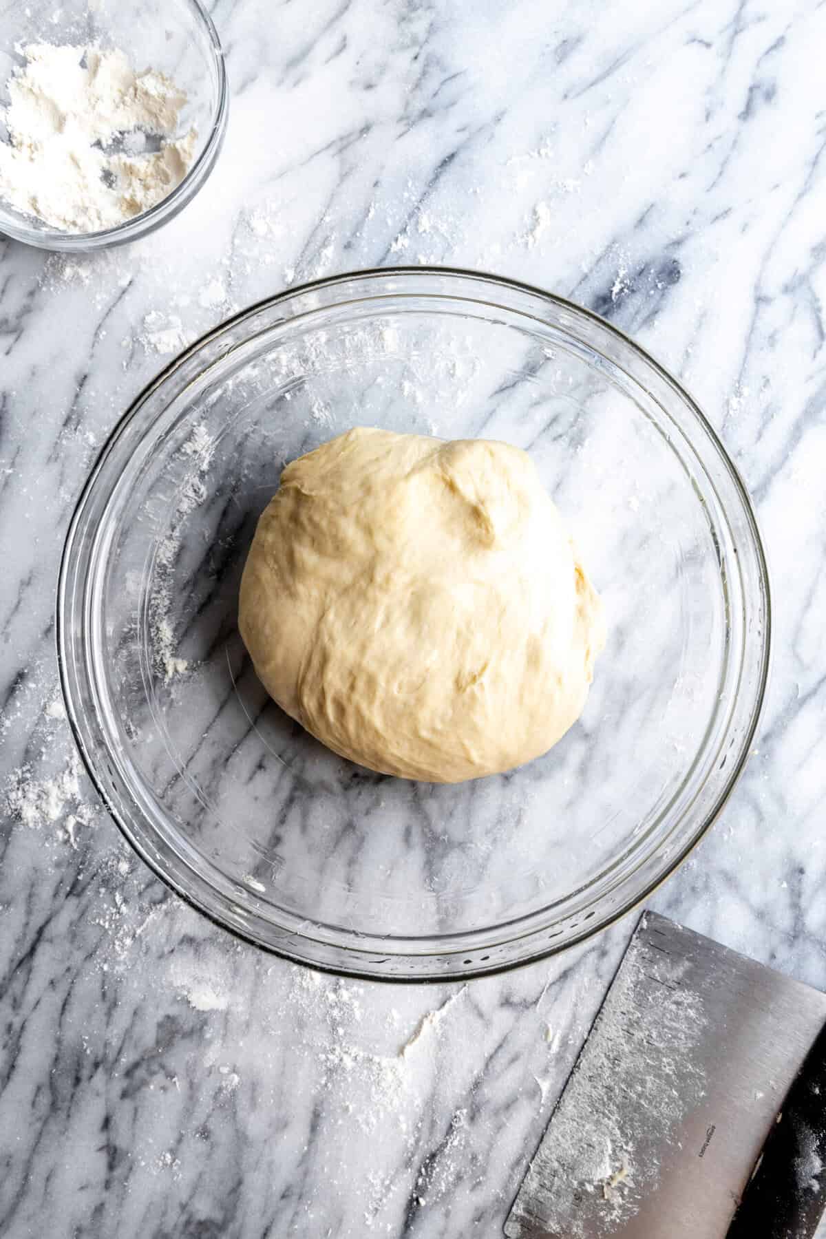 Pizza dough in a glass bowl and a bench scrape.
