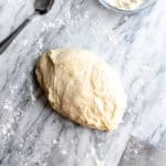 Pizza dough on a marble surface and a bowl of flour.