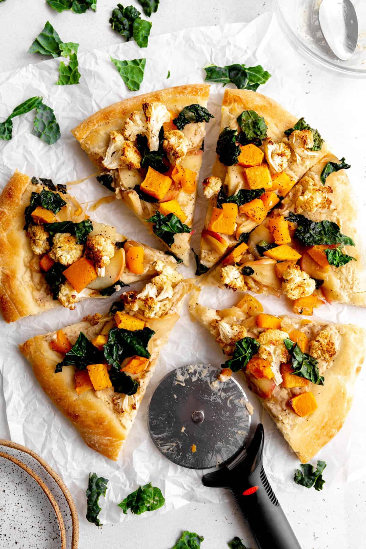 Roasted fall veggie pizza sliced on a white surface and a pizza cutter.