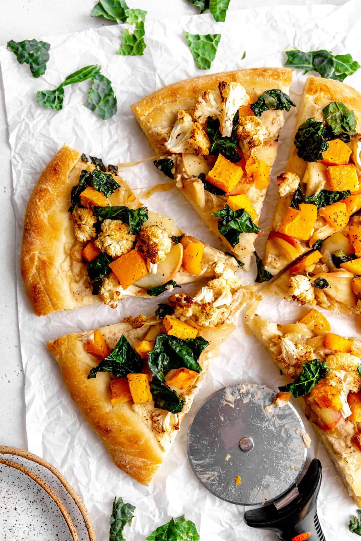 Roasted fall veggie pizza sliced on a white surface and a pizza cutter.