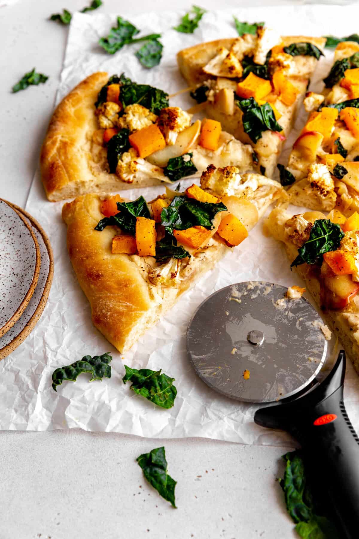 Slices of roasted veggie pizza on a white surface with a stack of plates.