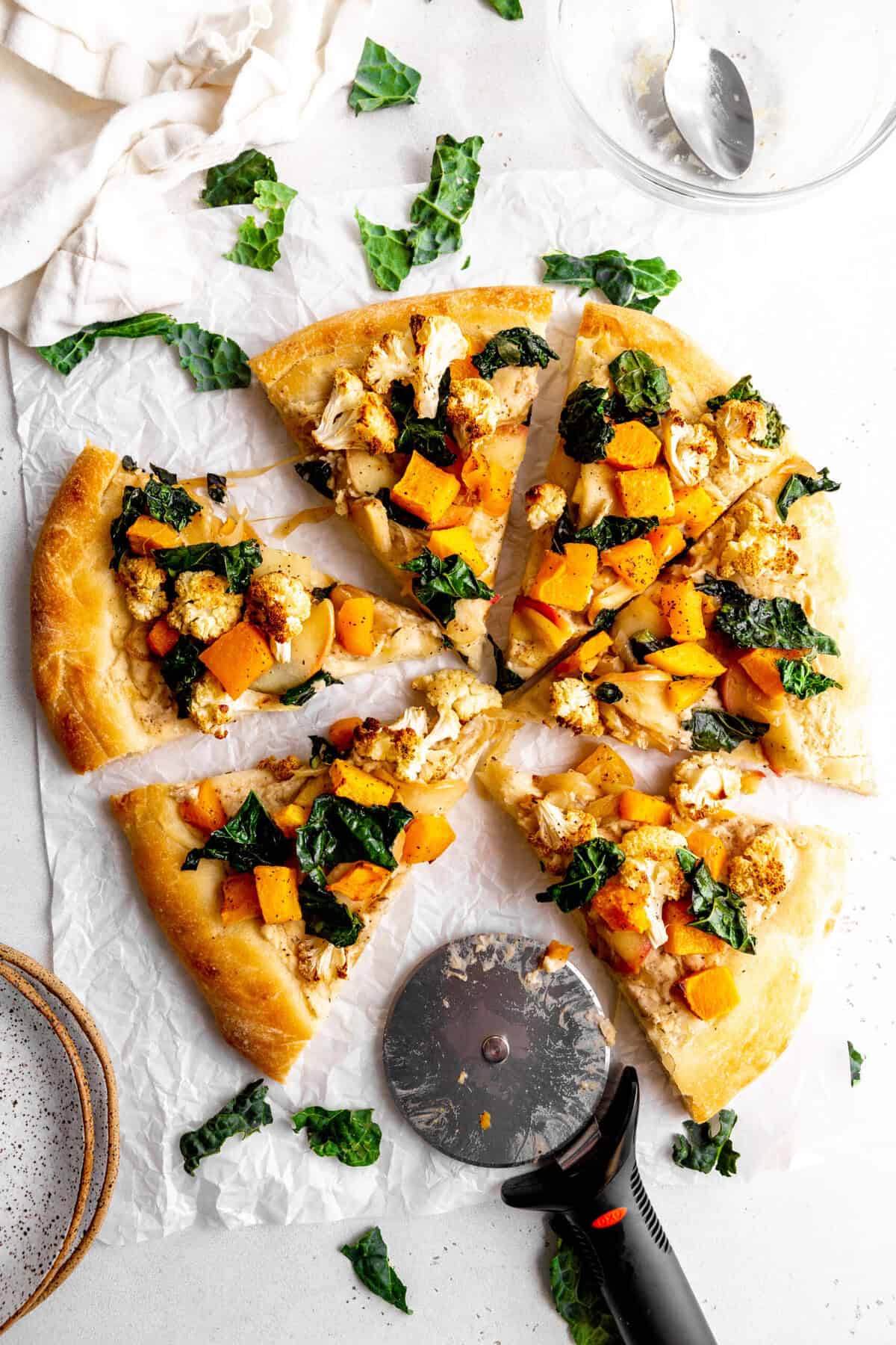 Roasted fall veggie pizza sliced on a white surface and a pizza cutter.