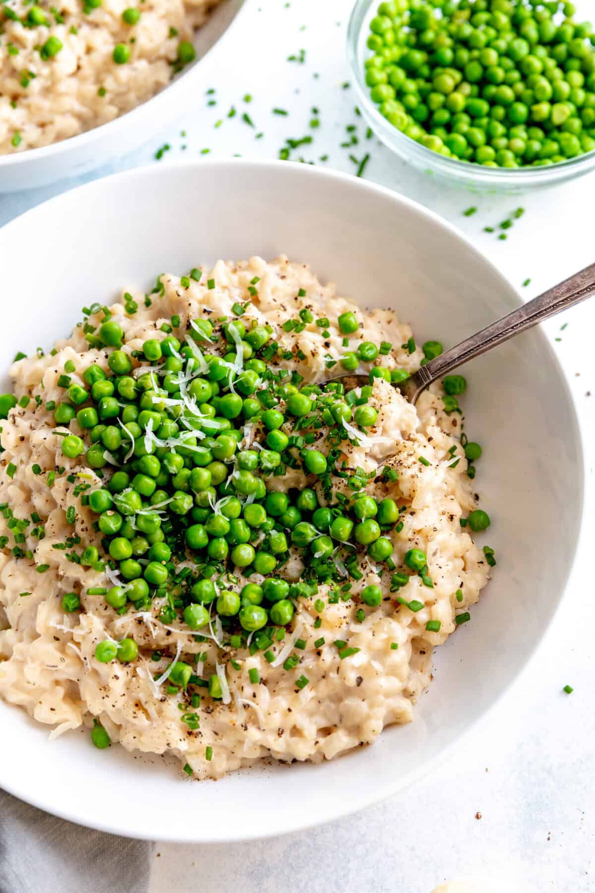 Spoon sticking in a bowl of risotto topped with green peas and a small bowl of peas.