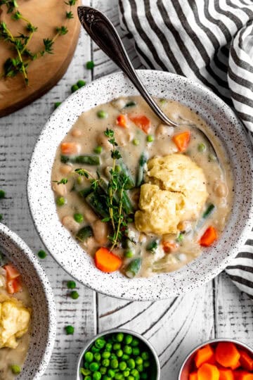 Spoon in a bowl of vegetable pot pie soup and small cups of peas and carrots.