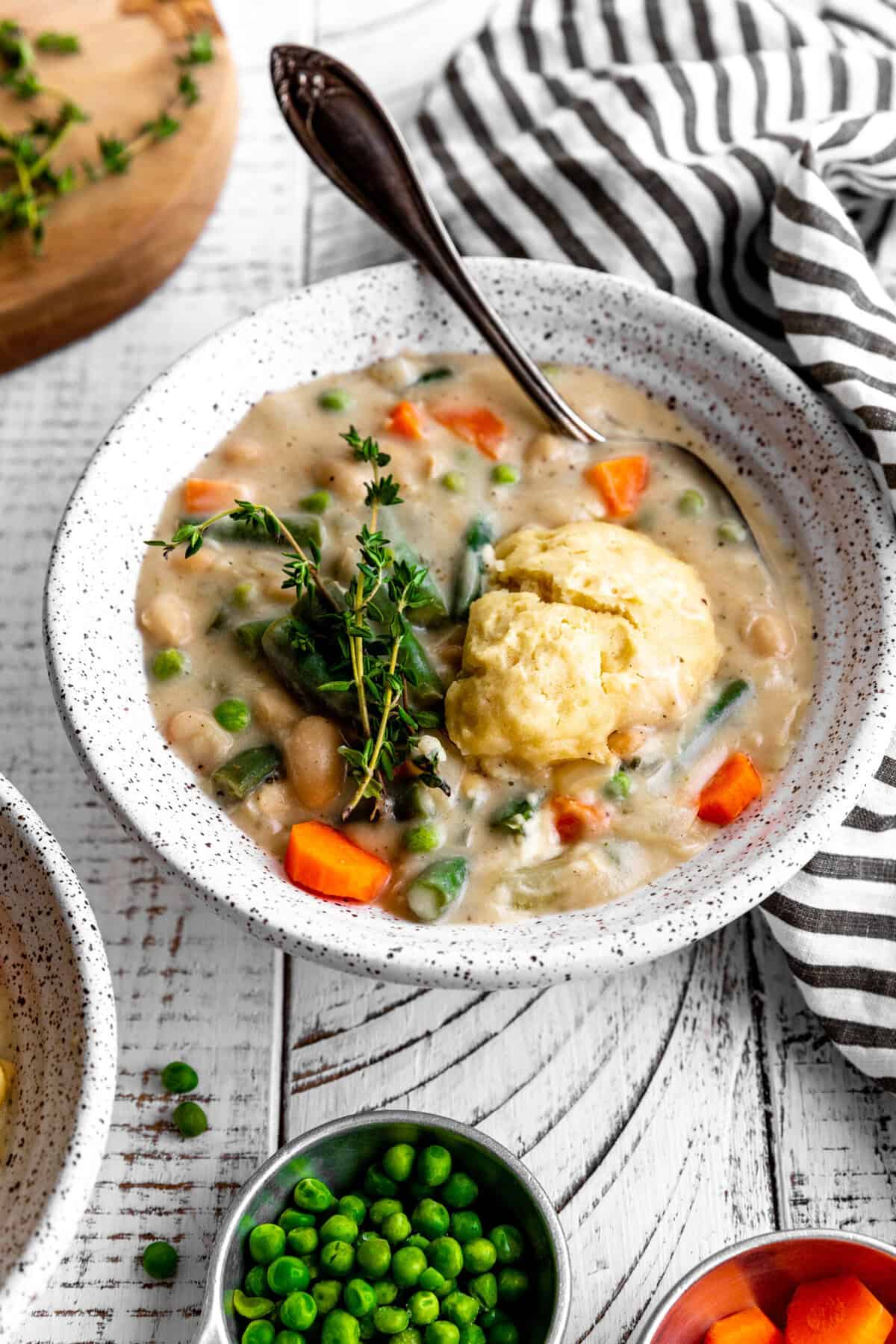 Spoon in a bowl of vegetable pot pie soup and small cups of peas and carrots.