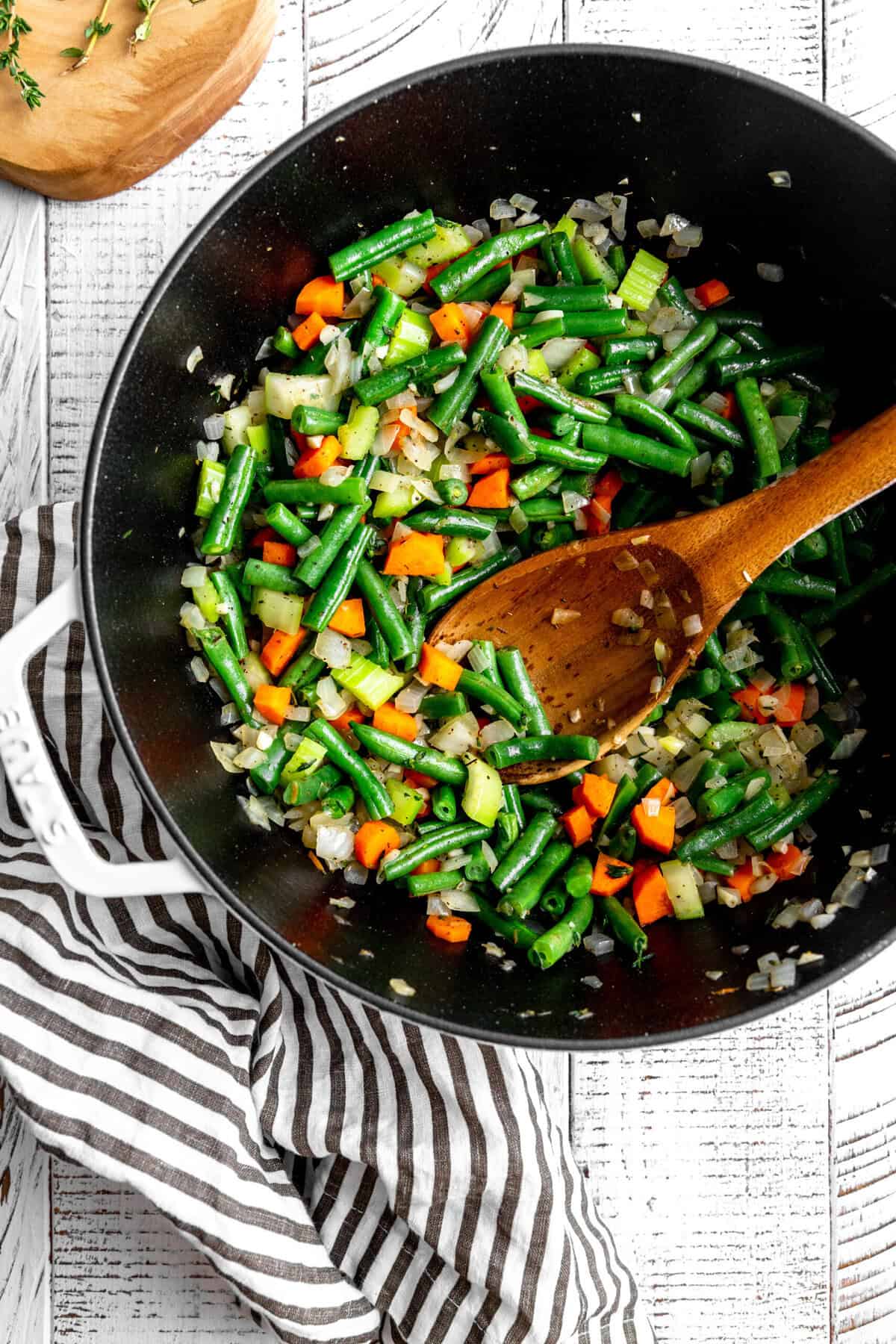 Sautéed onions, carrots, celery and green beans in a pot with a wooden spoon.