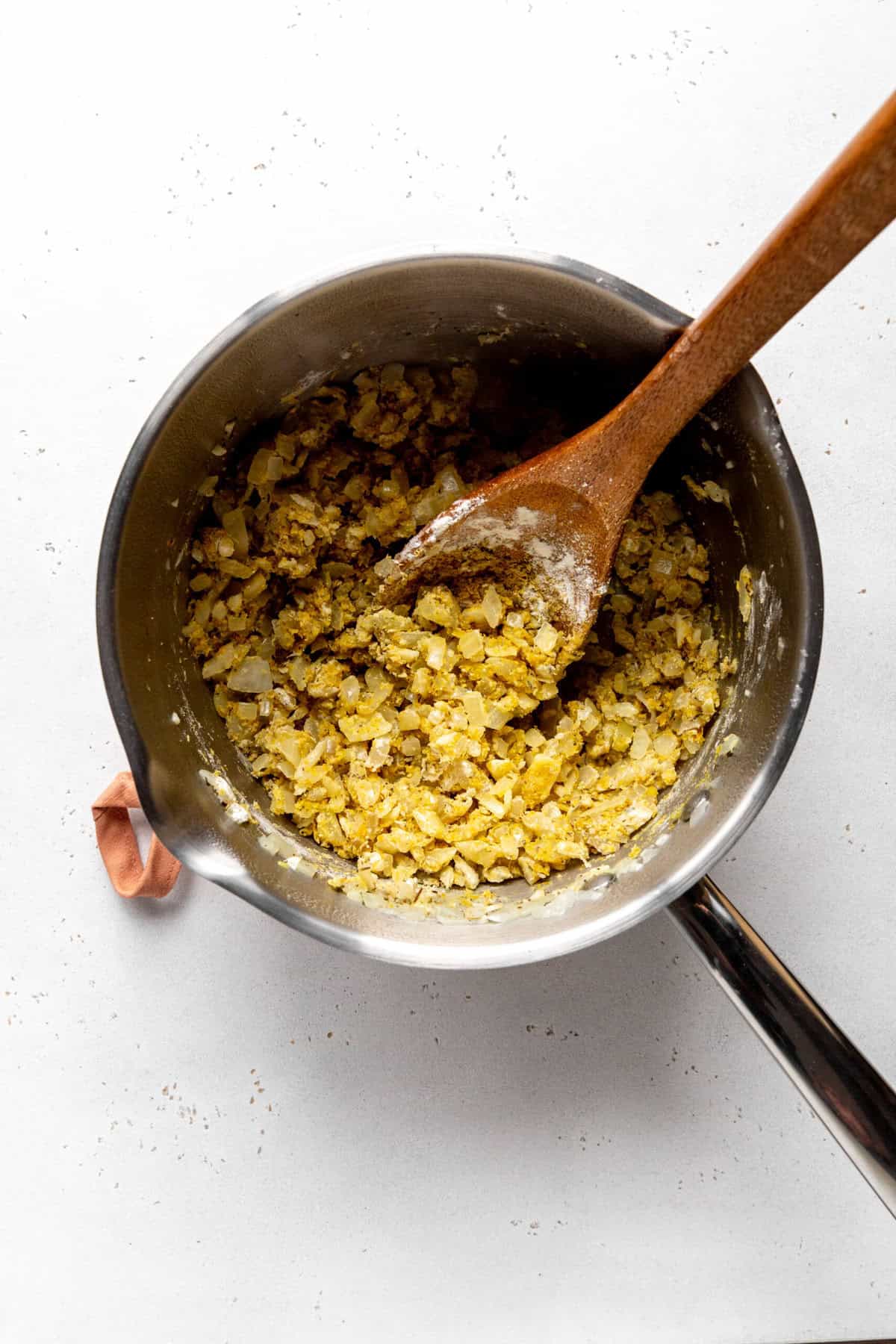 Sautéed onions covered in flour and nutritional yeast in a saucepan with a wooden spoon.