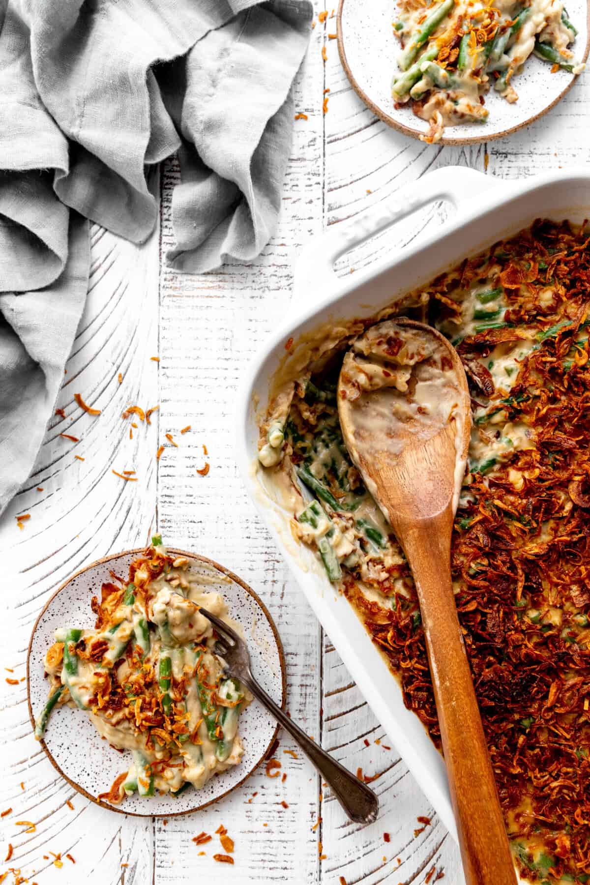 Dish of green bean casserole with a wooden spoon in it and two plates of casserole.
