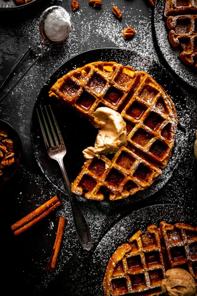 Pumpkin waffle with a section missing on a plate surrounded by cinnamon sticks.