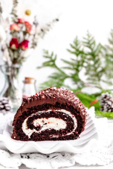 Peppermint yule log on a white plate, two pinecones and a vase of red roses.