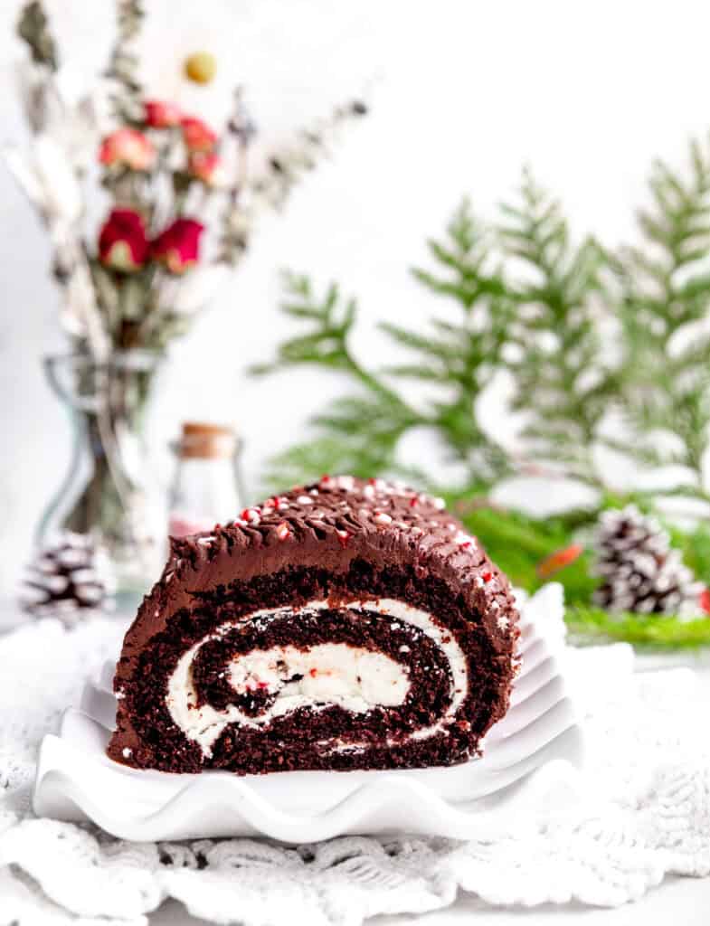 Peppermint yule log on a white plate, two pinecones and a vase of red roses.