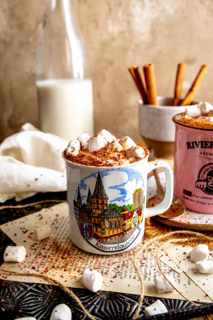 Two mugs of Mexican hot chocolate, a jug of milk and a cup of cinnamon sticks.