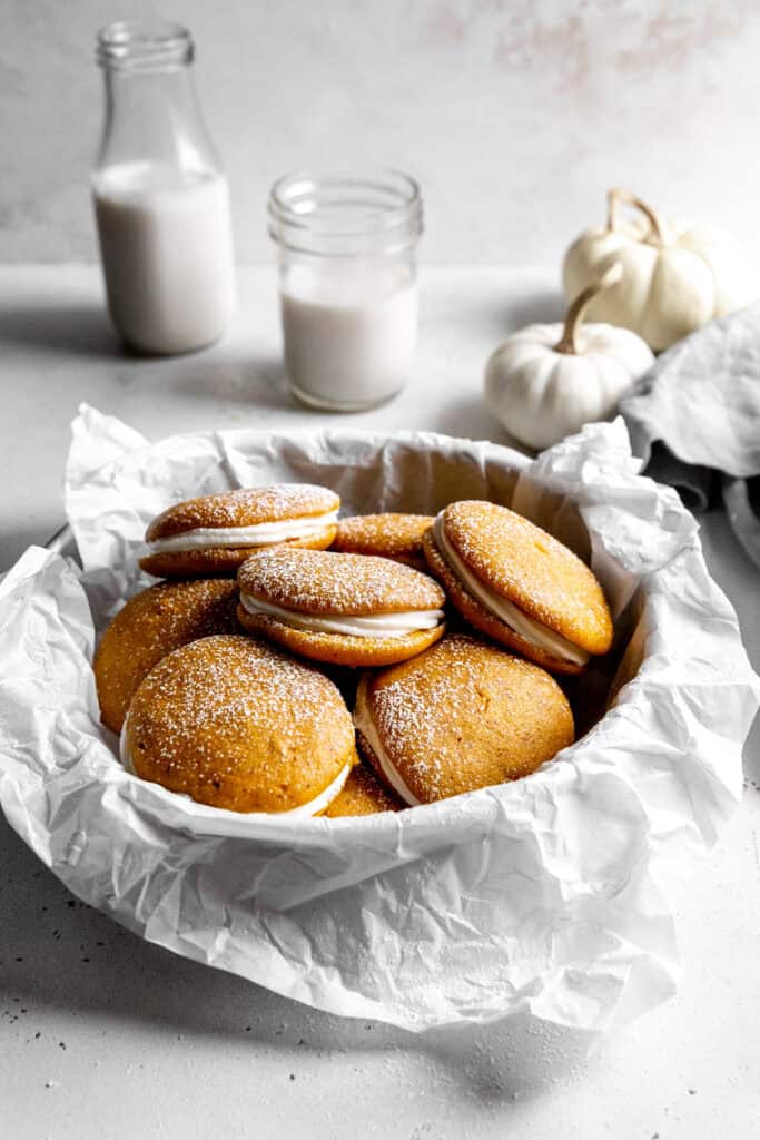 Bowl of pumpkin whoopie pies dusted with powdered sugar and a glass of milk.