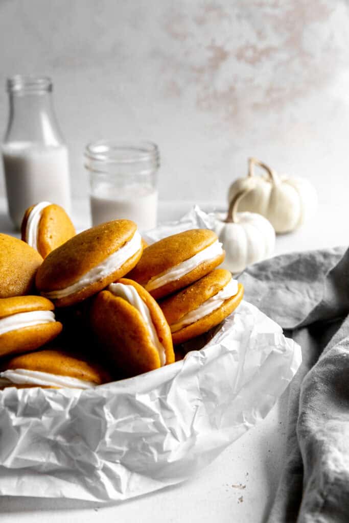 Bowl of stacked pumpkin whoopie pies, a linen napkin and two white pumpkins.