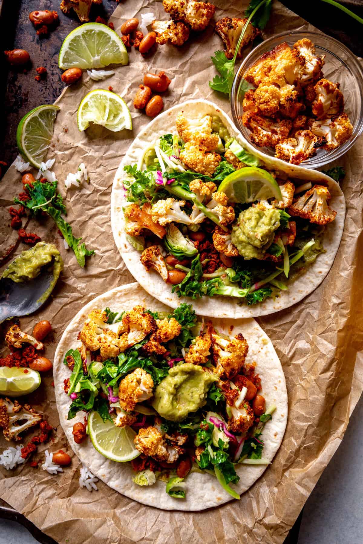 Cauliflower tacos on a baking sheet topped with guacamole and lime wedges.