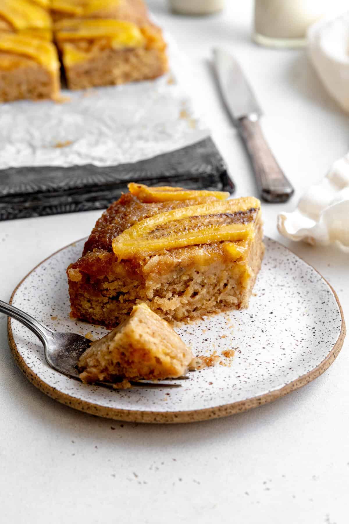 Fork cutting into a slice of banana upside down cake and a linen knife.