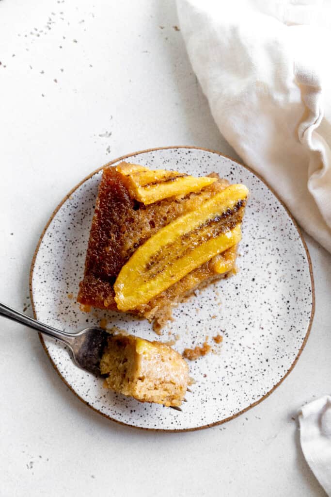 Fork cutting into a slice of banana upside down cake and a linen napkin.