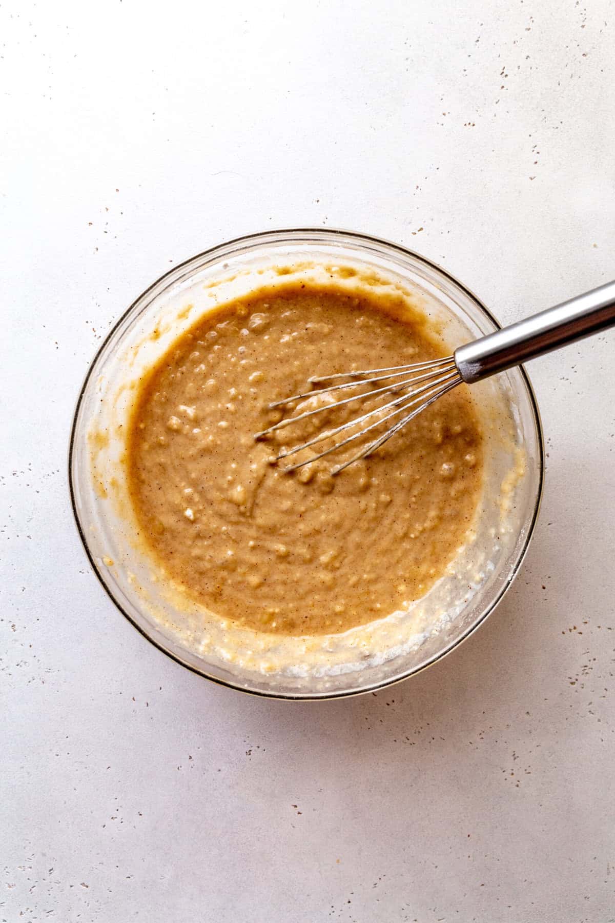 Glass bowl of banana cake batter and a wire whisk.