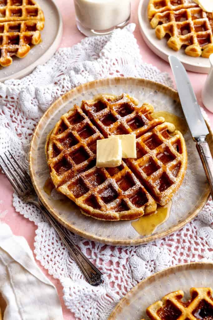 Fork and knife on a plate with a waffle covered in maple syrup and powdered sugar.