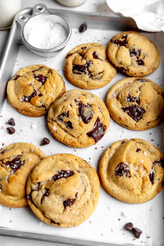 Chocolate chip cookies on a baking sheet with a cup of flaky salt.