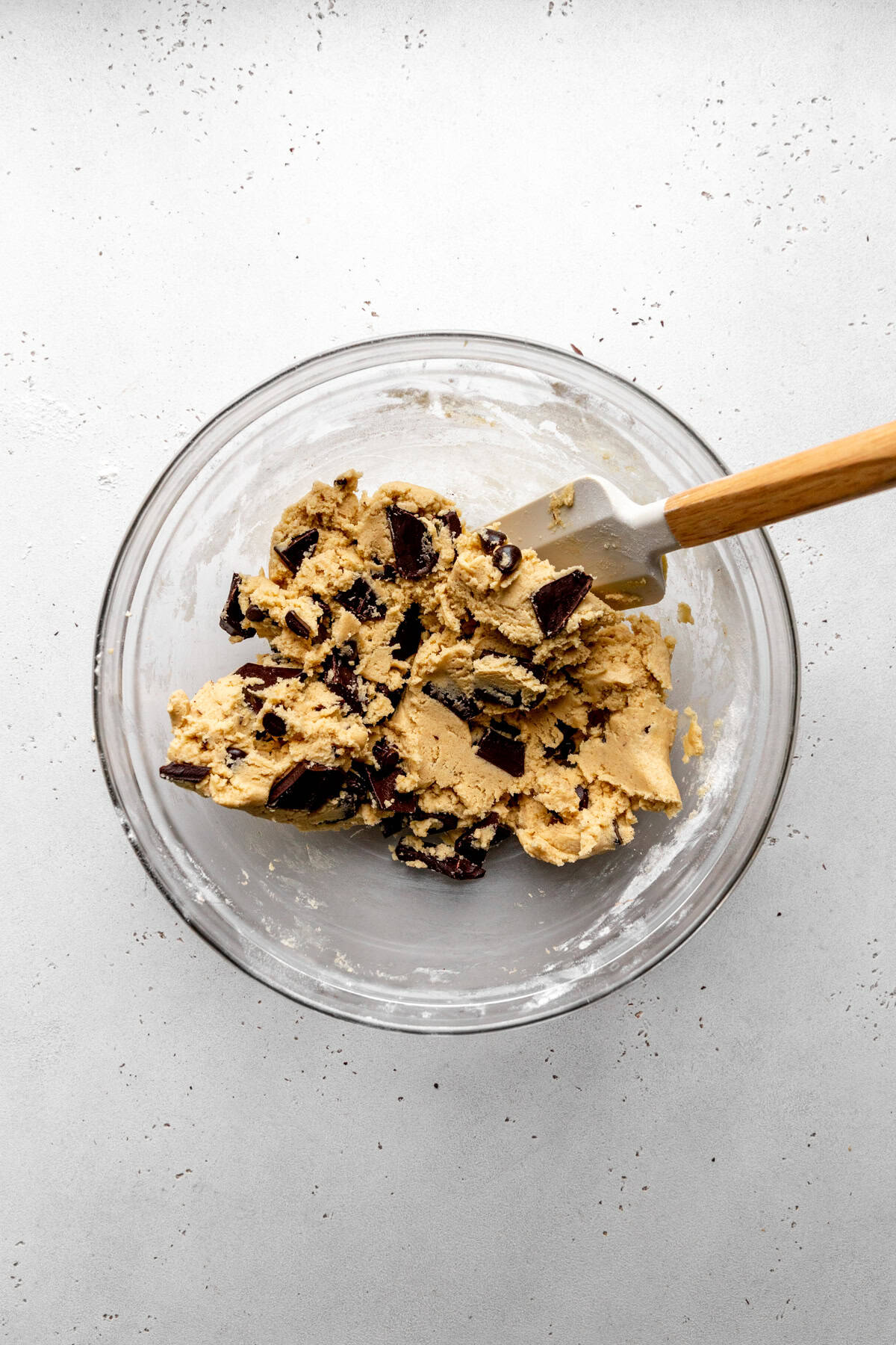 Chocolate chip cookie dough in a glass bowl with a rubber spatula.
