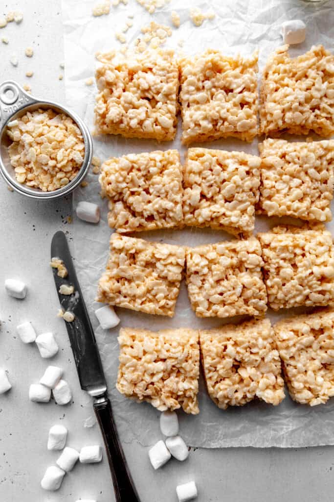 Rice krispie treats, a cup of rice krispies, scattered marshmallows and a knife.