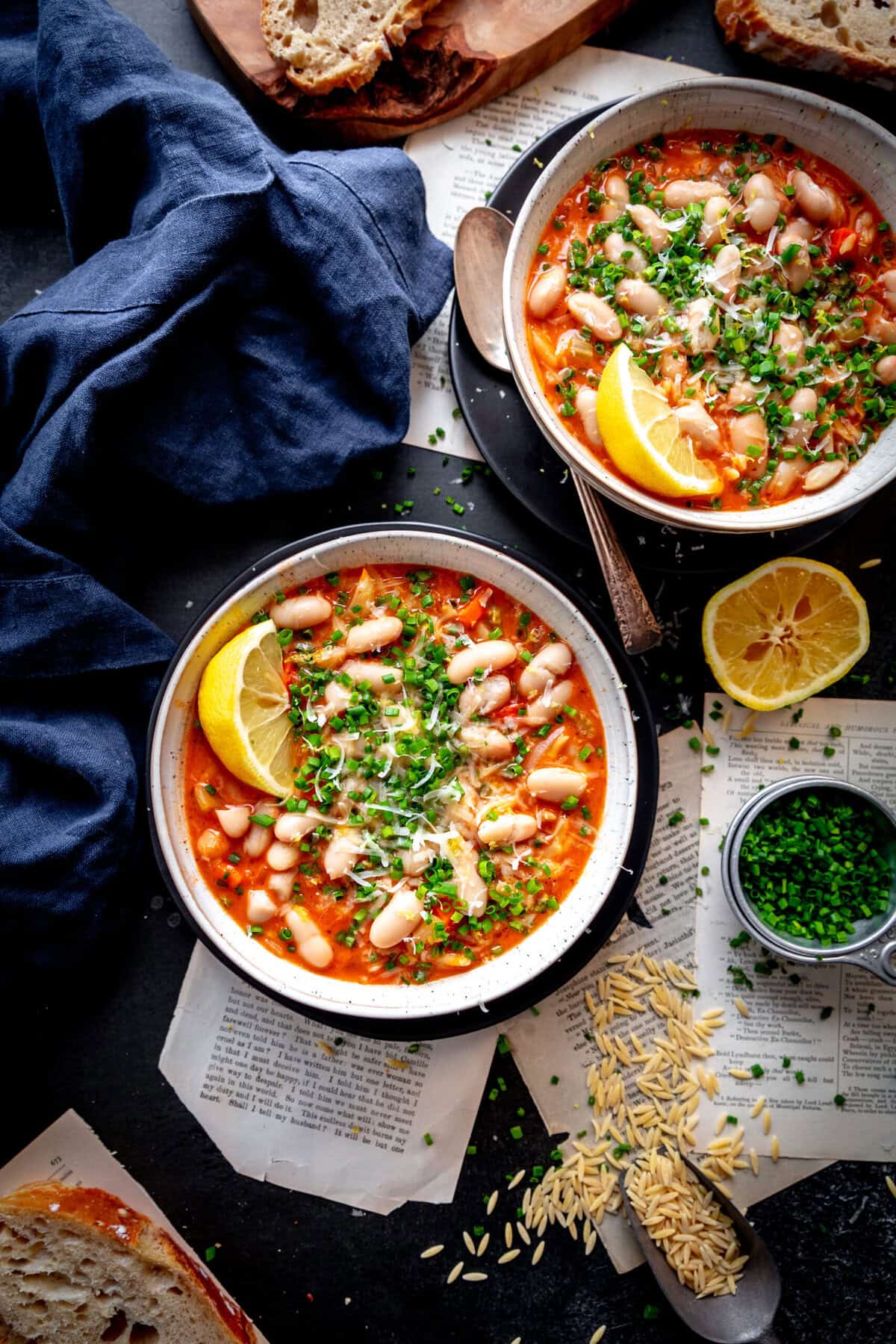 Two bowls of orzo tomato soup, slices of bread, and a cup of chopped chives.