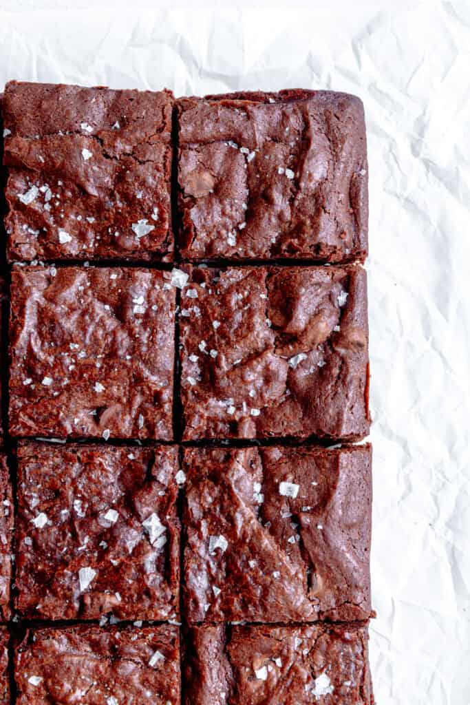 Vegan brownies topped with flaky sea salt on white parchment paper.