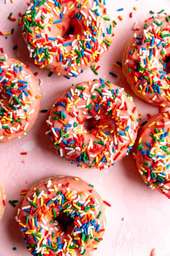 Baked donuts topped with pink icing and rainbow sprinkles on a pink surface.