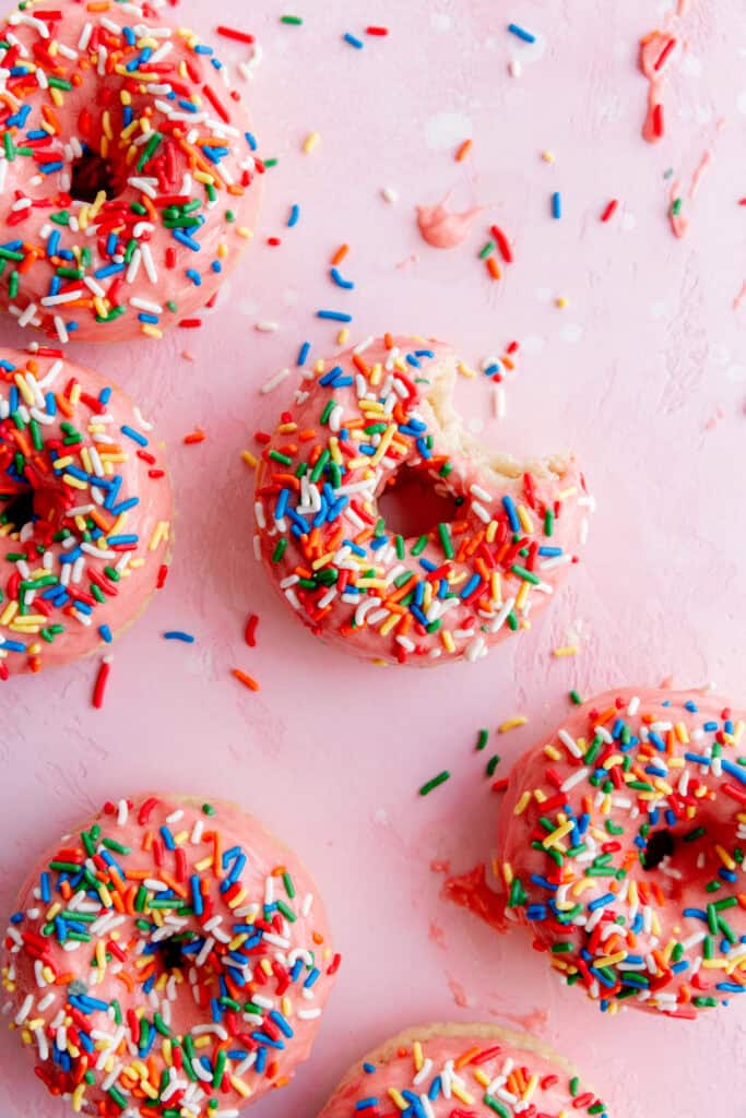 Baked donuts with strawberry icing and rainbow sprinkles on a pink surface.