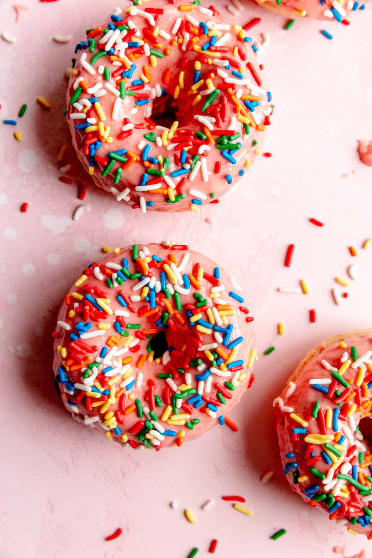 Baked, pink sprinkle donuts on a pink surface covered in rainbow sprinkles.
