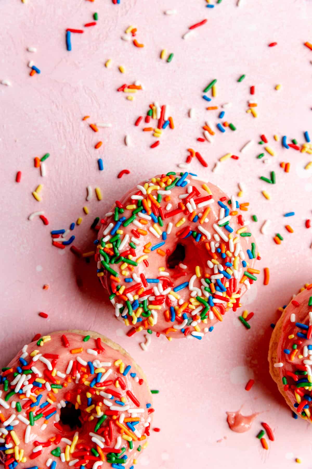 Pink sprinkle donuts on a pink surface covered in rainbow sprinkles.