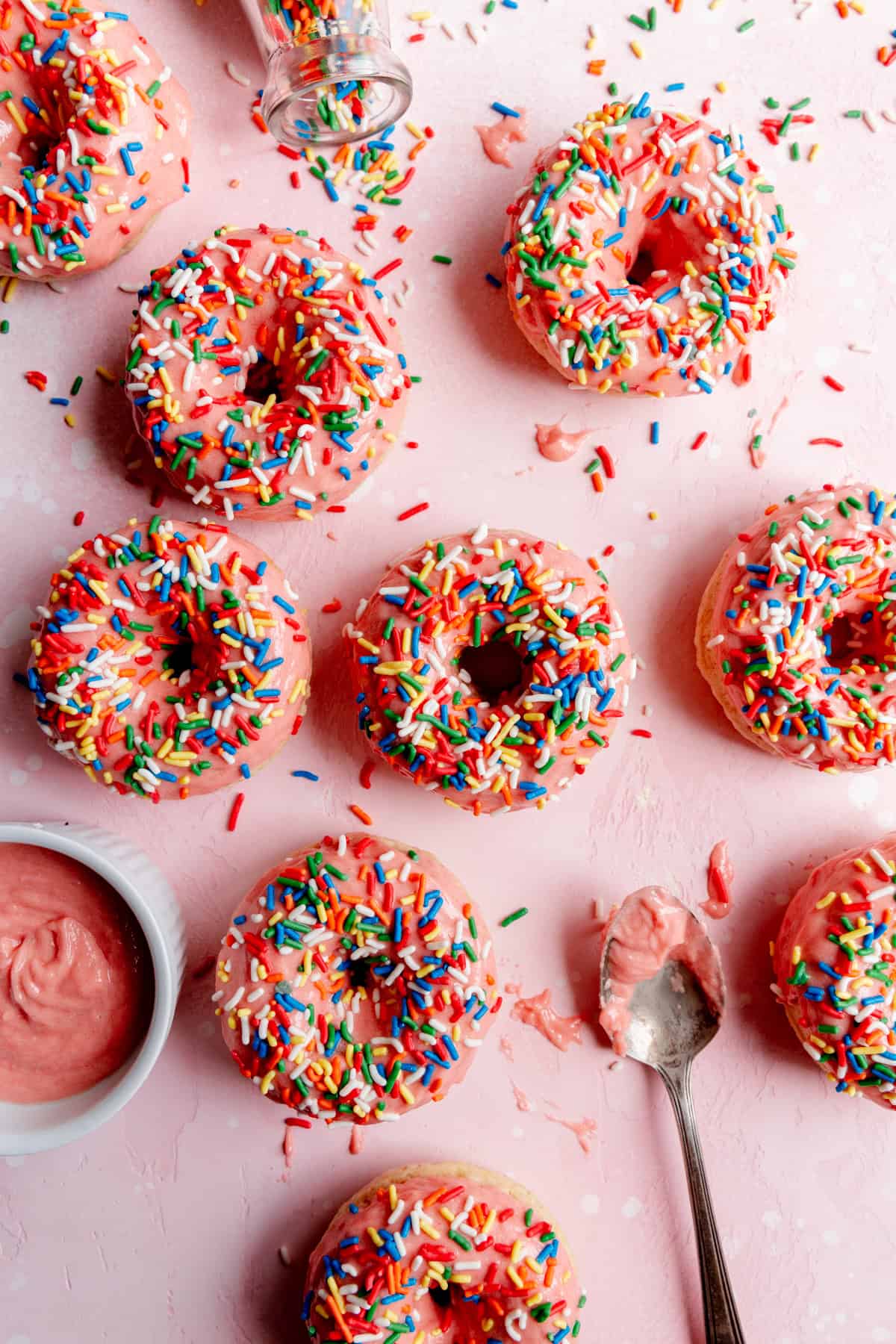 Pink sprinkle donuts, spilled rainbow sprinkles, and a bowl of pink frosting.