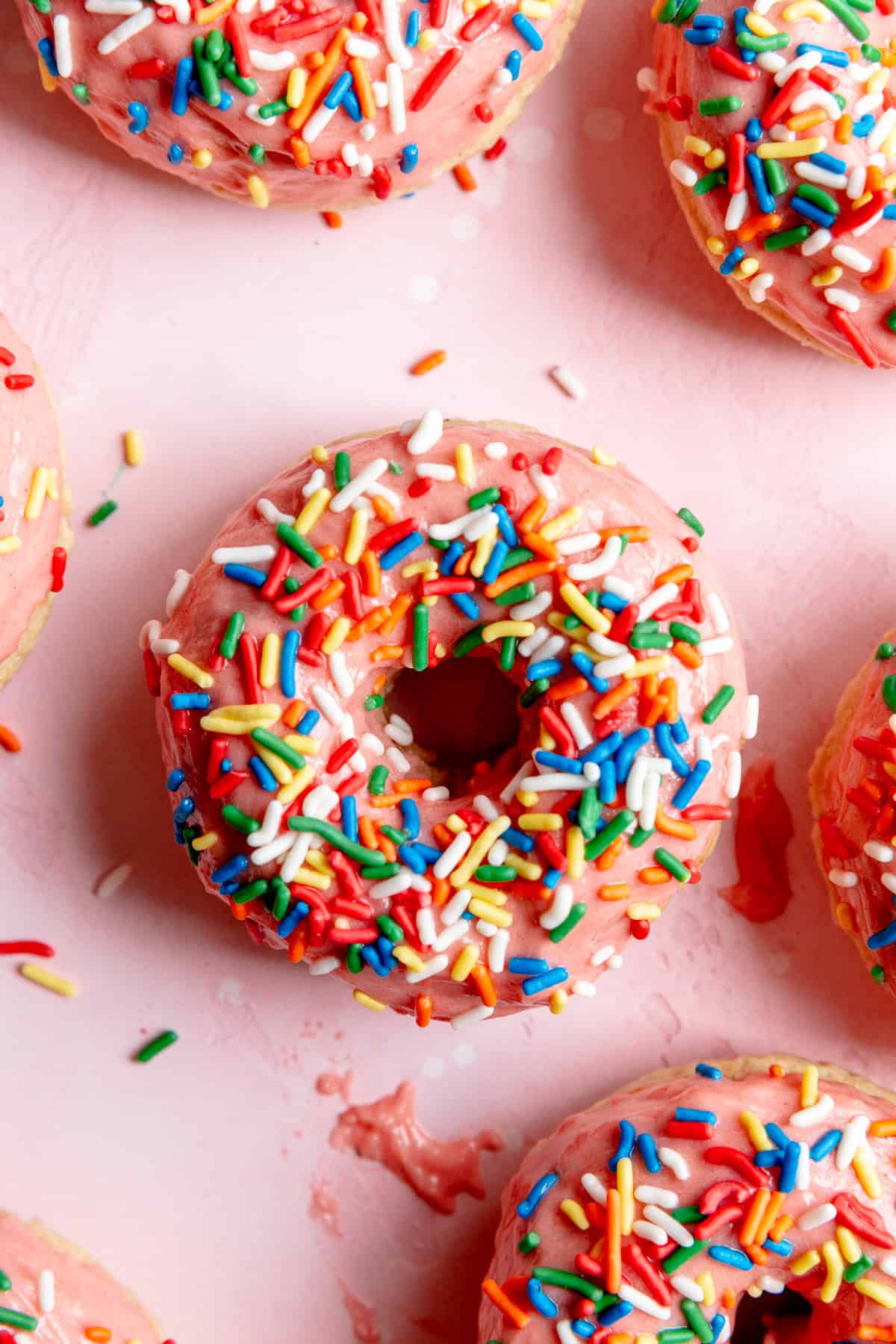 Baked donut with pink icing and rainbow sprinkles on a pink surface.