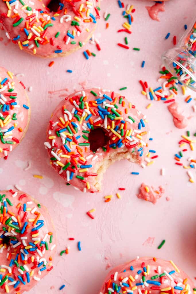 Baked donut topped with pink icing and rainbow sprinkles with a bite taken out of it.
