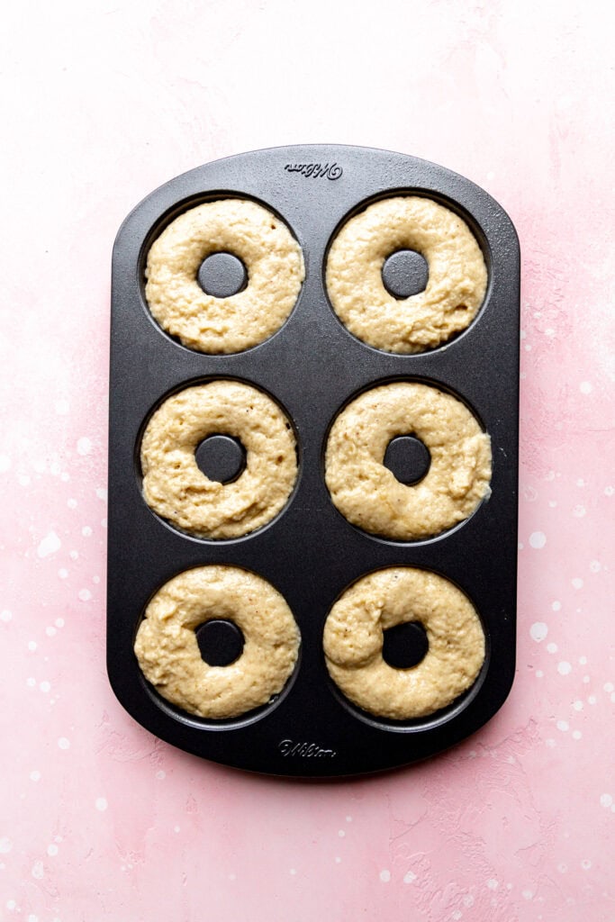 Unbaked donuts in a donut pan on a pink surface.