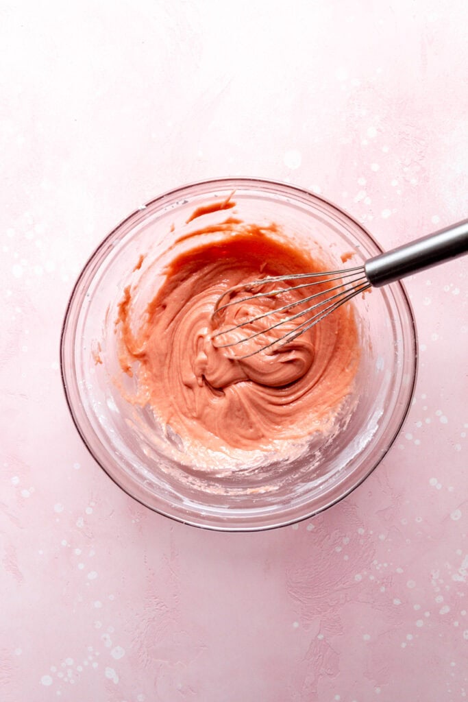 Strawberry icing in a glass bowl with a whisk.