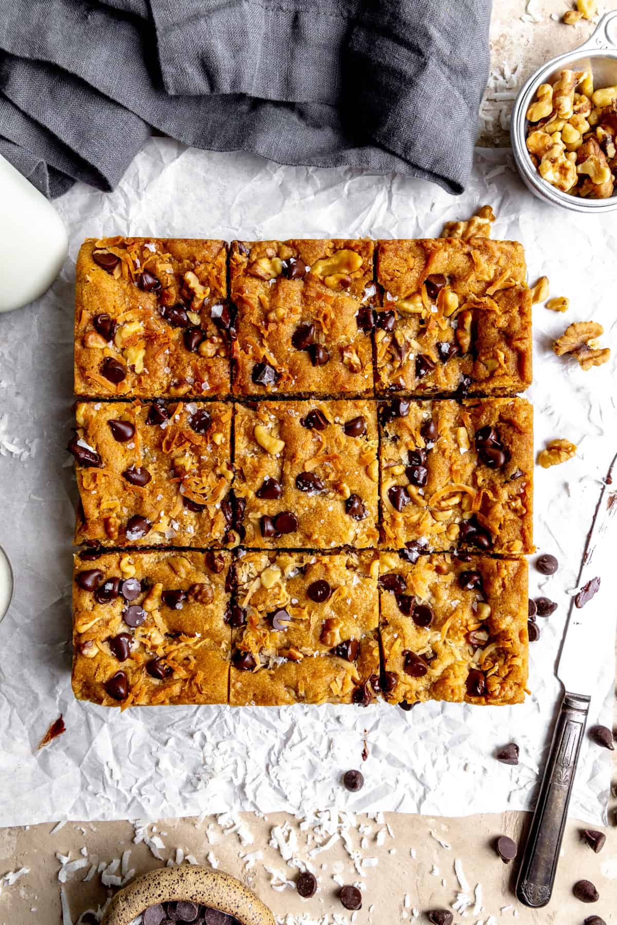 Vegan blondies, a knife, a linen napkin, a glass of milk and a cup of walnuts.